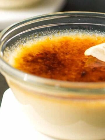 A glass bowl of crème brûlée with a caramelized sugar top, garnished with a lemon slice. It sits on a white coaster, accompanied by a spoon on a dark surface. Another blurred bowl is in the background.