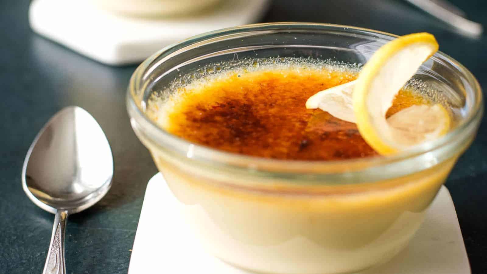 A glass bowl of crème brûlée with a caramelized sugar top, garnished with a lemon slice. It sits on a white coaster, accompanied by a spoon on a dark surface. Another blurred bowl is in the background.