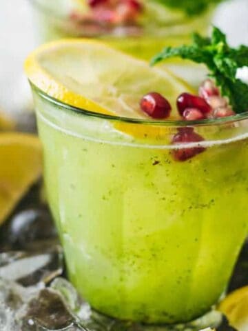 A glass of bright green mojito garnished with a lemon slice, pomegranate seeds, and mint leaves. Several decorative glasses are in the background. Lemon wedges and ice cubes surround the glass on the table.
