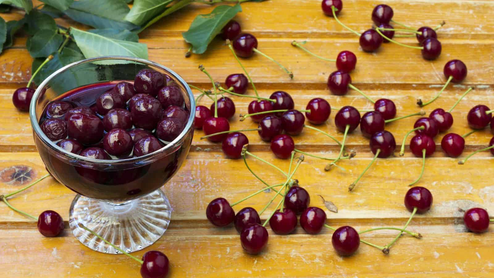 A glass bowl filled with dark cherries in juice is placed on a wooden surface. Several whole cherries with stems are scattered around, along with green leaves in the background. The wood has a yellowish tint and a weathered texture.