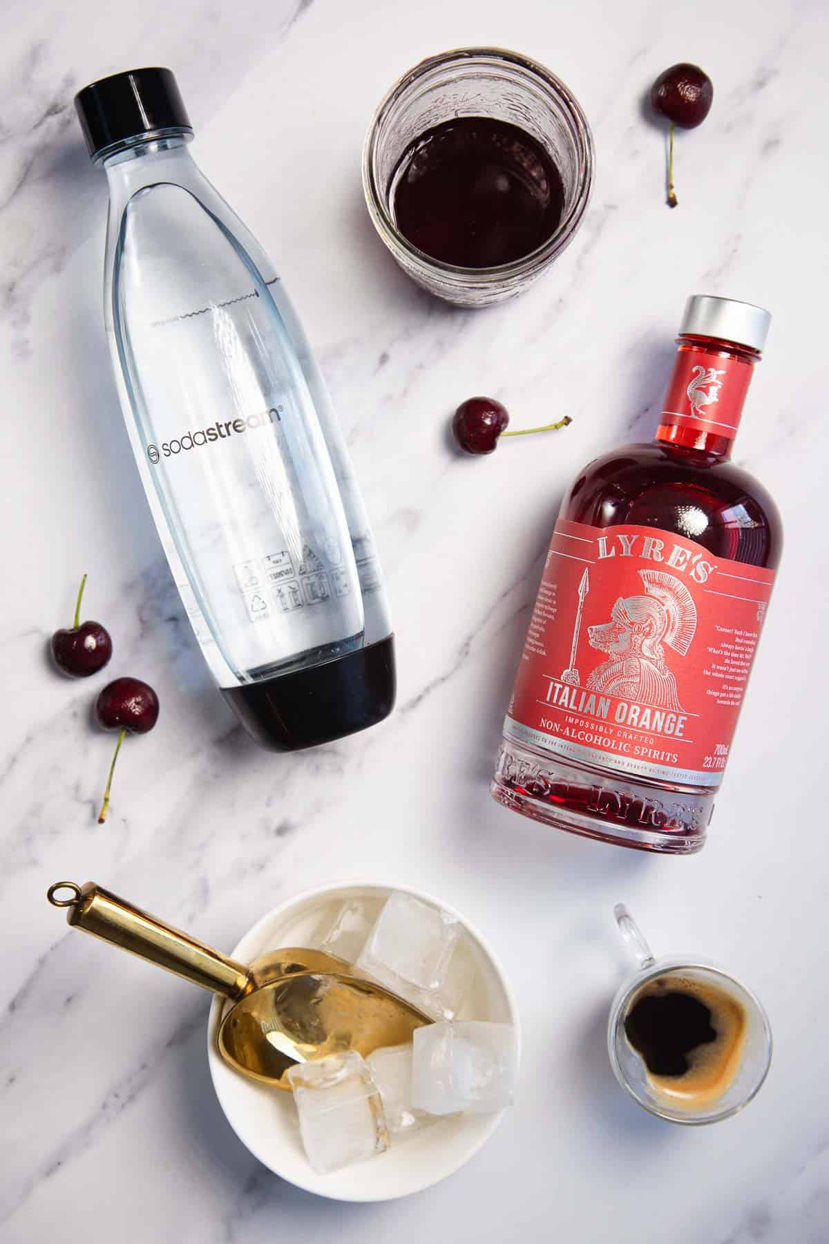 A bottle of sparkling water, a bottle of Lyre's Italian Orange, a glass with dark liquid, scattered cherries, a bowl with ice cubes and a gold spoon, and a small cup of espresso are arranged on a marble surface.
