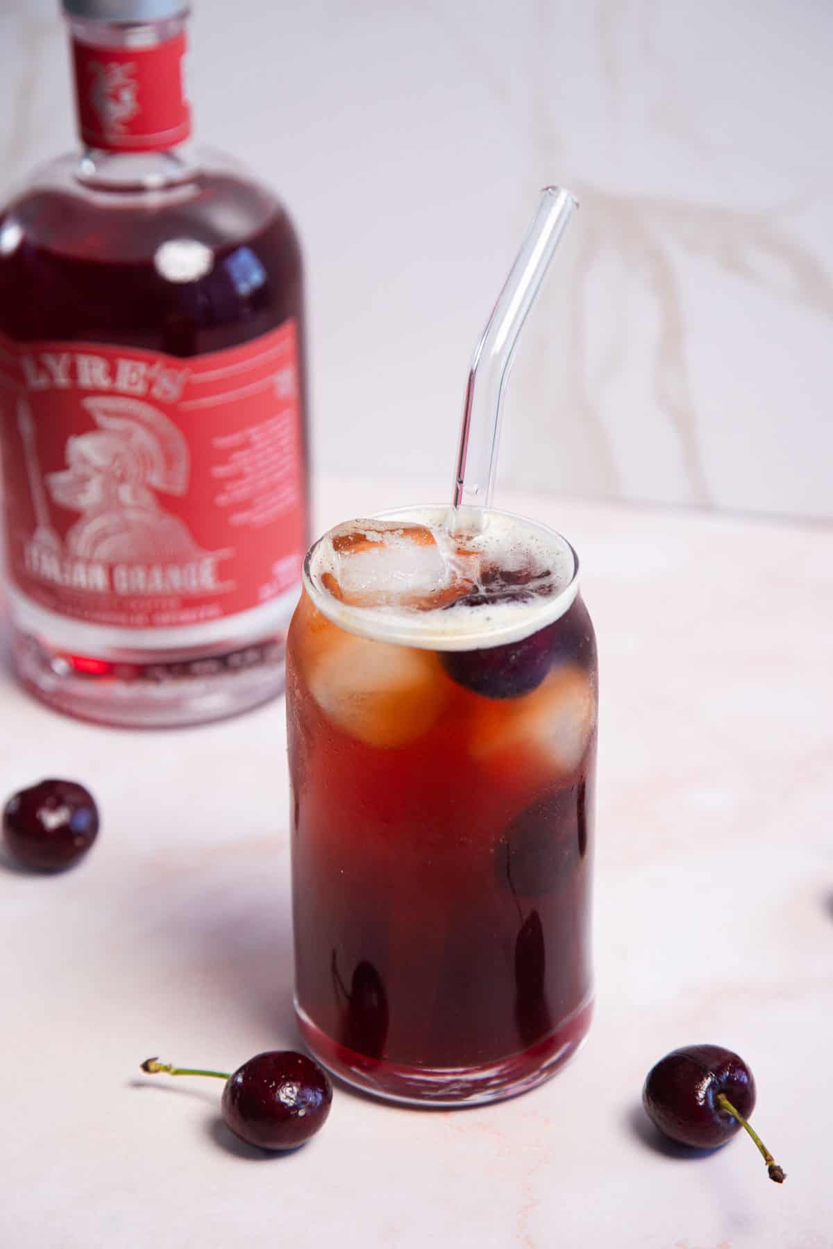 A glass of dark iced drink with a clear straw sits on a light surface, surrounded by cherries. In the background, there is a bottle of Lyre's Italian Orange.
