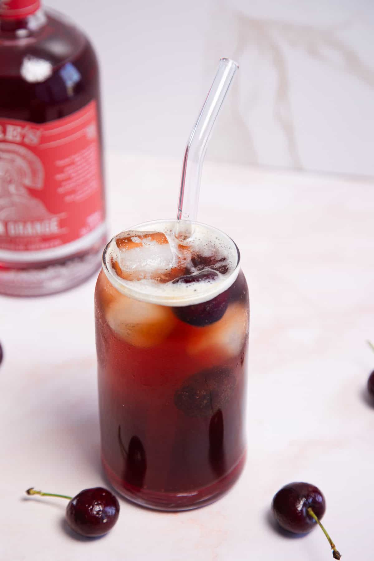 A glass filled with iced dark liquid and a clear straw, garnished with cherries, stands on a light pink surface. A bottle with a red label is partially visible in the background, along with a few scattered cherries.
