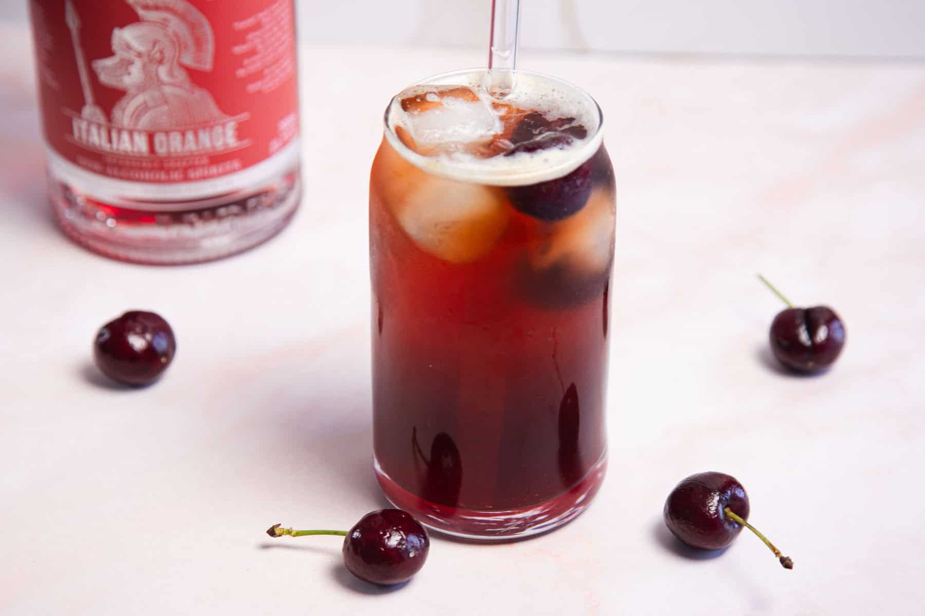 A tall glass filled with a reddish-brown beverage and ice sits on a light surface. A straw is placed in the drink. Fresh cherries are scattered around the base of the glass. A bottle with a red label is partially visible in the background.