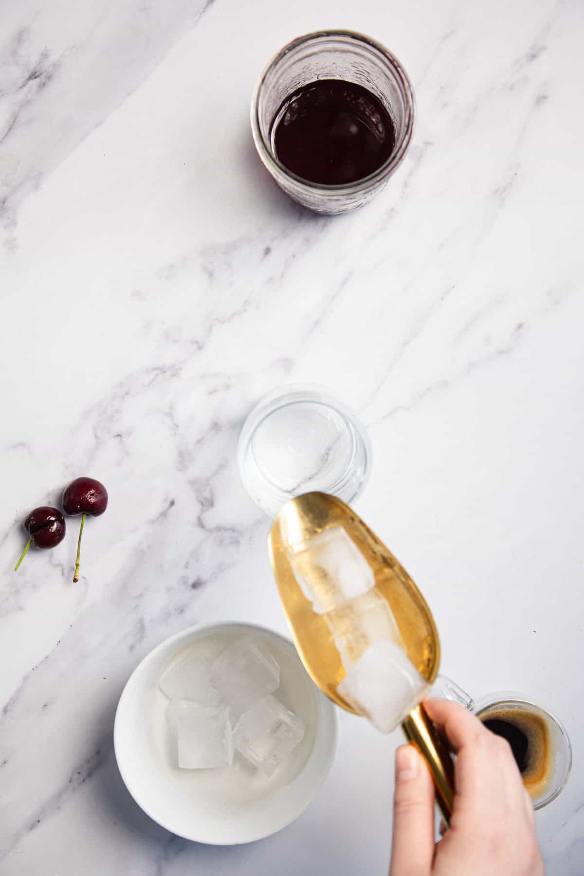 A hand is placing ice cubes into a glass with a gold-colored scoop. Nearby, there is an empty white bowl with more ice cubes, two cherries, a glass filled with dark liquid, and a small cup of espresso on a marble surface.