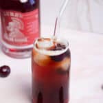 A glass filled with a dark cherry-colored drink and ice cubes, with a clear straw. Fresh cherries are scattered nearby. In the background, there is a blurry bottle with a red label. The surface is white.