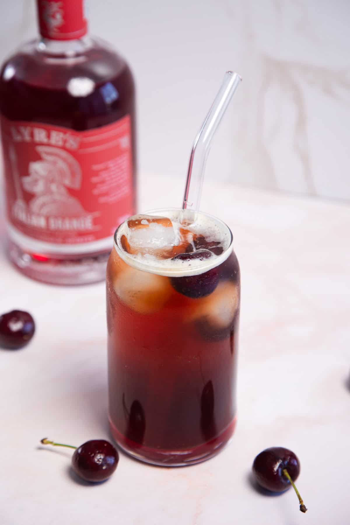 A glass filled with a dark cherry-colored drink and ice cubes, with a clear straw. Fresh cherries are scattered nearby. In the background, there is a blurry bottle with a red label. The surface is white.