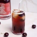 A glass filled with a dark iced beverage, topped with two cherries and ice cubes, with a straw inserted. A bottle with a red label is partially visible in the background. Several cherries are scattered on the light-colored surface.