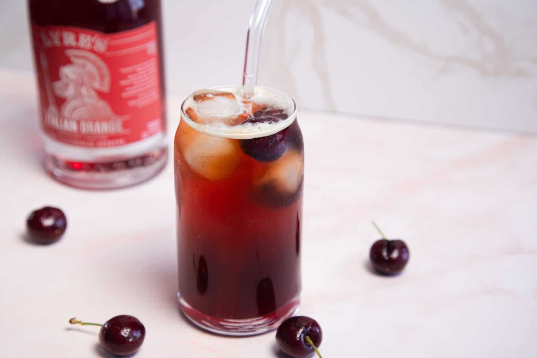 A glass filled with a dark red beverage, ice cubes, and a straw sits on a light surface. Several cherries are scattered around the glass. In the background, there is a bottle with a red label.