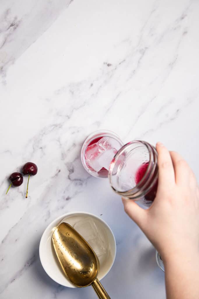 A hand is pouring a red drink from a jar into a glass with ice, on a marble surface. Nearby, there are two cherries and a bowl with a large gold spoon inside.