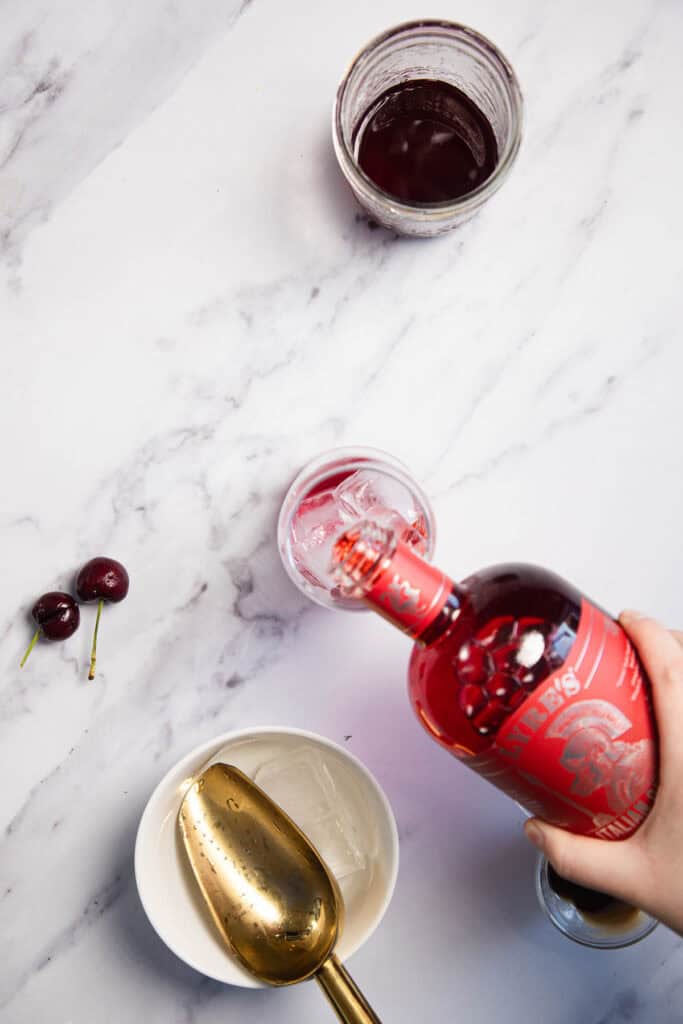 A hand pours red liquid from a bottle labeled "Mister's" into a glass with ice cubes. A mixing glass with dark liquid, a bowl with a gold scoop, and two cherries on the marble surface are also visible.