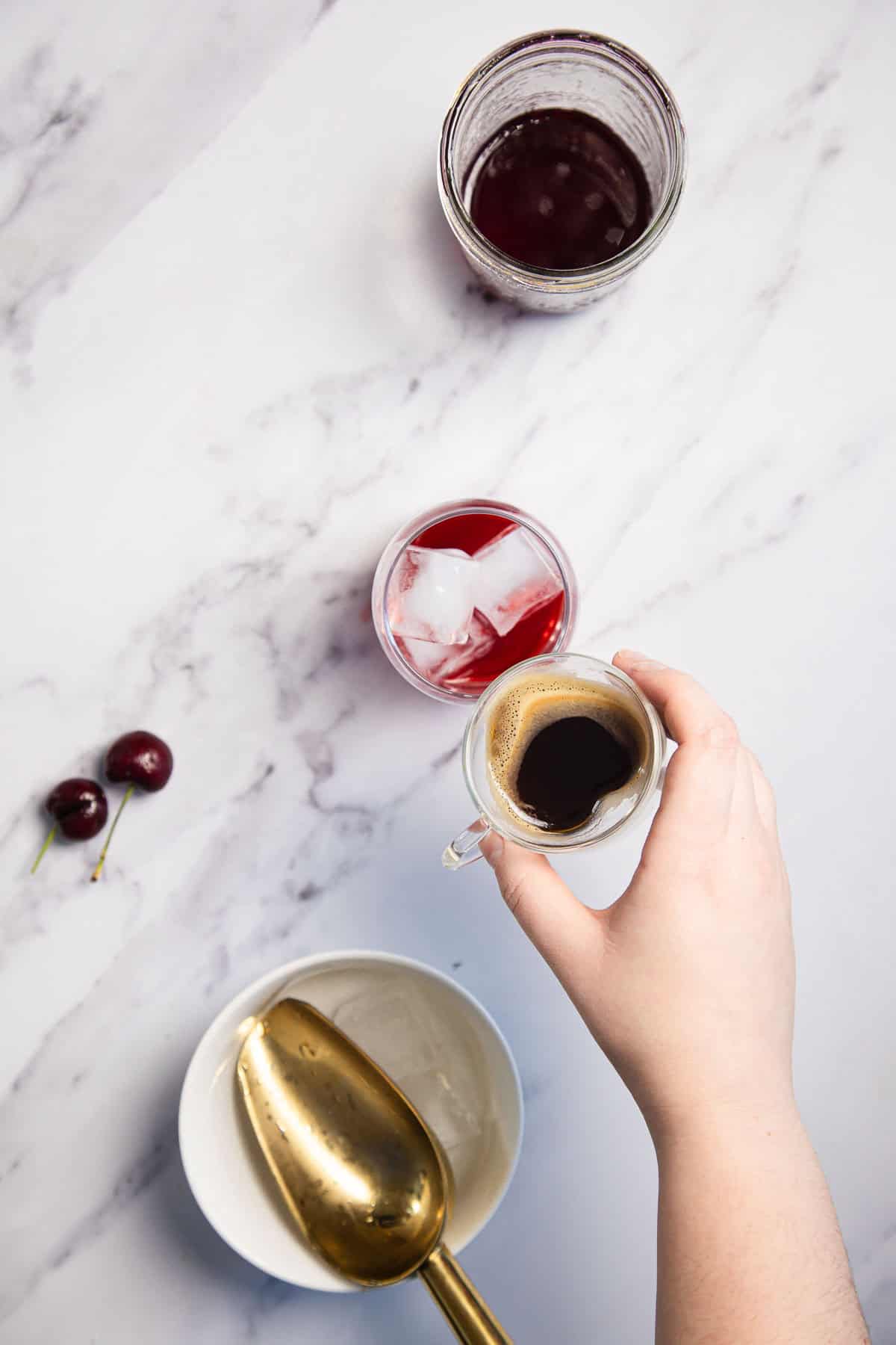 A hand holding a small cup of espresso above a drink with ice cubes and red liquid on a marble surface. Beside it, there's a glass with more red liquid, two cherries, and a bowl with a golden scoop.