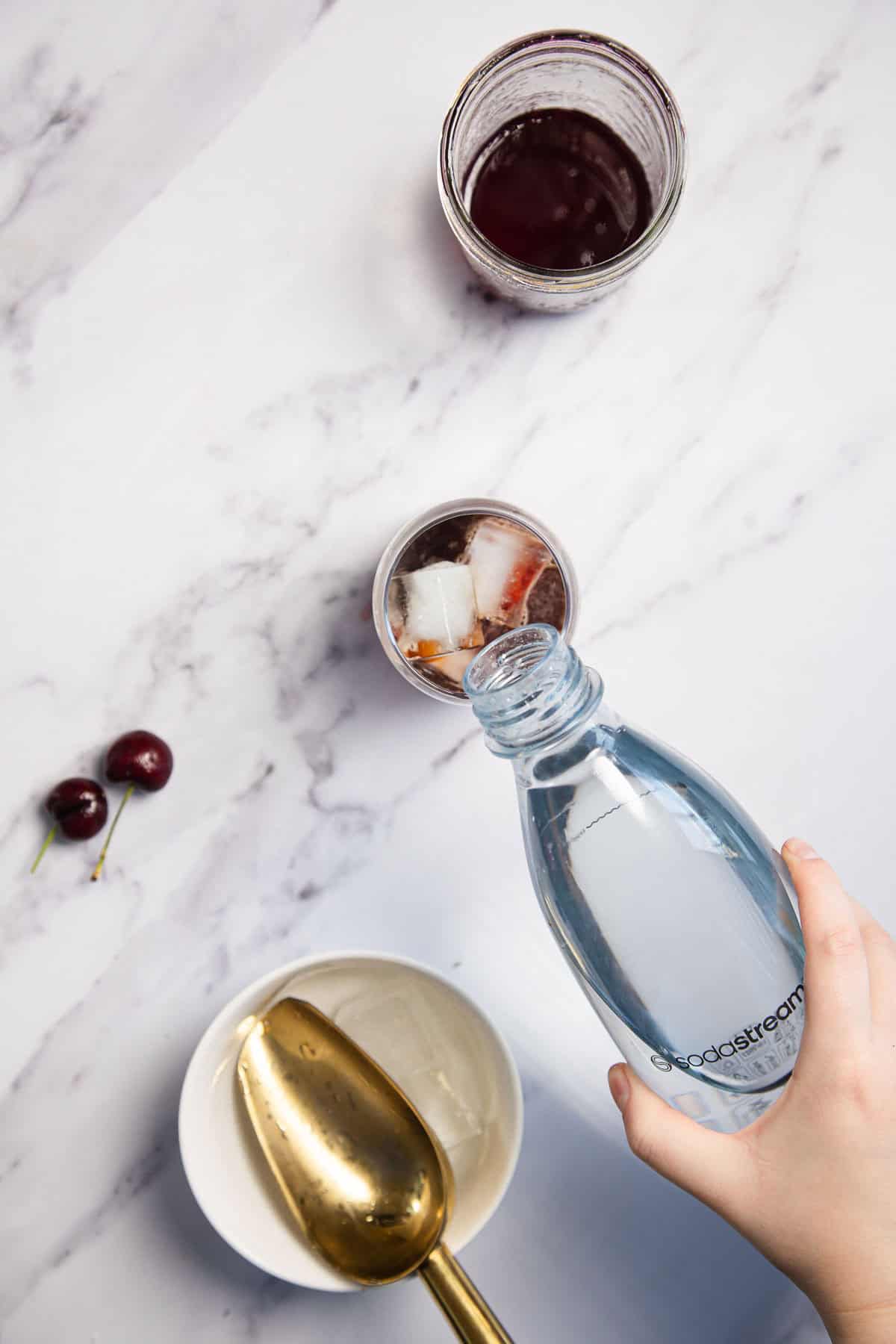 A hand pours water from a bottle labeled "Sodastream" into a glass with ice. Nearby are a jar of dark liquid, two cherries, and a white bowl with a gold-colored scoop. Everything is set on a white marble surface.