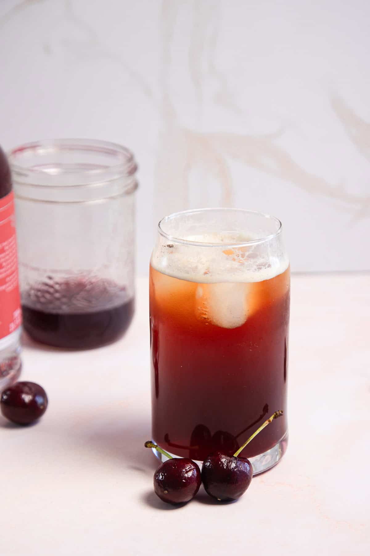 A glass of dark cherry americano fizz mocktail with foam, ice, and two cherries; jar and bottle in the background.