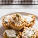 A plate with four small tarts topped with whipped cream, a sprinkle of cinnamon, and pumpkin seeds. A wire cooling rack with more tarts is visible in the background. The tarts rest on a marble surface.