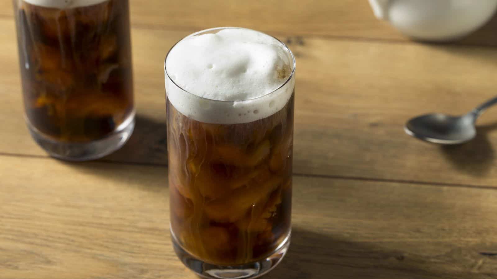 Two tall glasses filled with iced coffee topped with frothy foam are on a wooden table. A spoon is nearby, and a white pitcher is partially visible in the background.