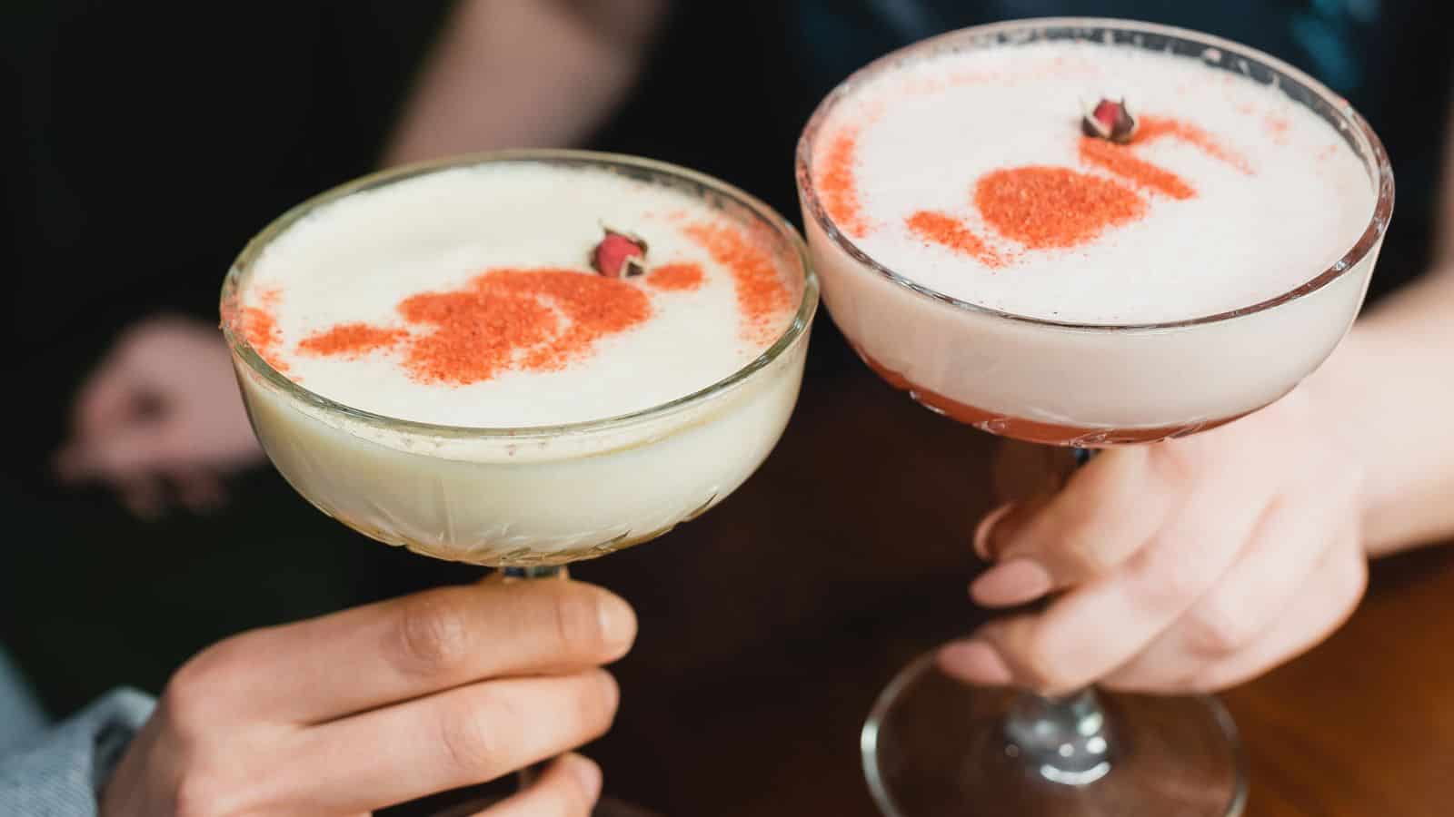 Two hands hold cocktail glasses filled with frothy drinks. Each glass is garnished with red sprinkles and small decorative elements on top. The glasses have a classic coupe shape and are raised in a toasting gesture.