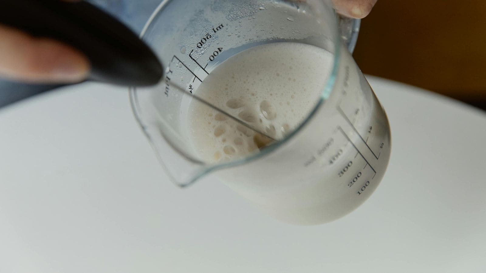 A person is frothing milk in a measuring cup using a handheld frother. The liquid is bubbling and filling part of the cup, which has measurement markings visible on the side. The scene is set against a plain, light-colored background.