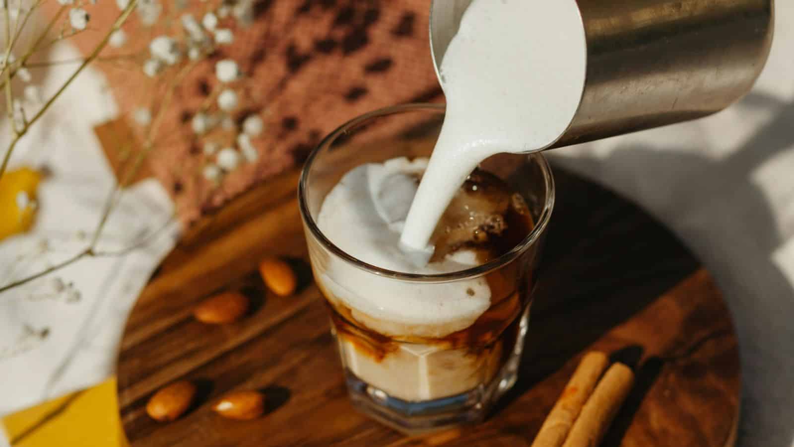 A glass of iced coffee is being topped with frothy milk poured from a metal pitcher. The drink sits on a wooden board, surrounded by almonds and two cinnamon sticks. Delicate white flowers are visible in the background.