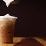 A plastic cup filled with iced coffee sits on a wooden table against a dark background. Foam is being poured onto the drink, creating a frothy top layer.