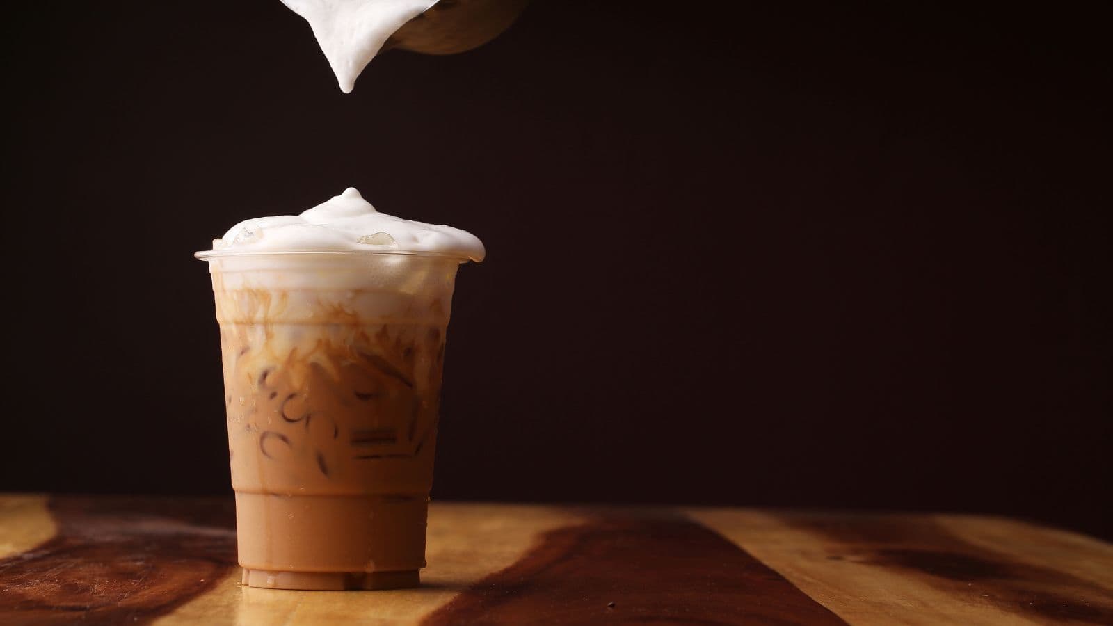 A plastic cup filled with iced coffee sits on a wooden table against a dark background. Foam is being poured onto the drink, creating a frothy top layer.
