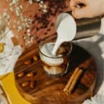 A hand is pouring milk foam from a metal jug into a glass of iced coffee on a wooden tray. The tray also holds almonds and cinnamon sticks. There are yellow books and white flowers in the background on a light fabric.