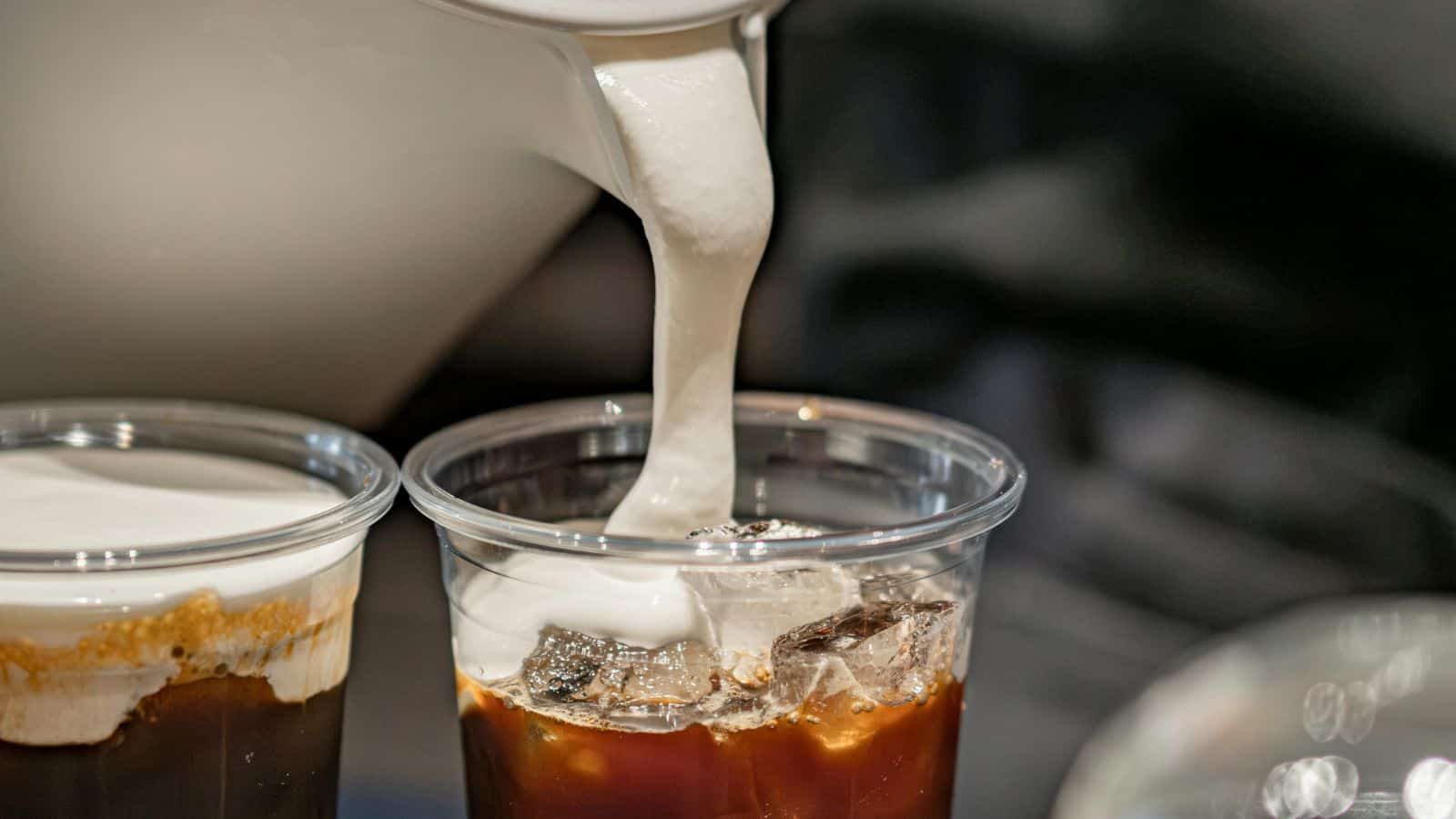 Close-up of a hand pouring thick white cream into a plastic cup filled with iced coffee, perfect for achieving that coffee shop cold foam at home. Another cup with iced coffee and a layer of cream on top is partially visible next to it.