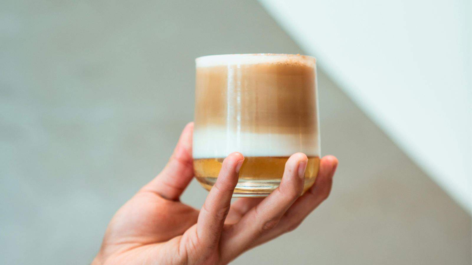 A hand holds a glass filled with a layered beverage, showcasing the perfect blend of brown, white, and cream hues. Soft lighting highlights the drink's elegance, making it easy to achieve that coffee shop cold foam aesthetic at home. The background remains artfully out of focus.