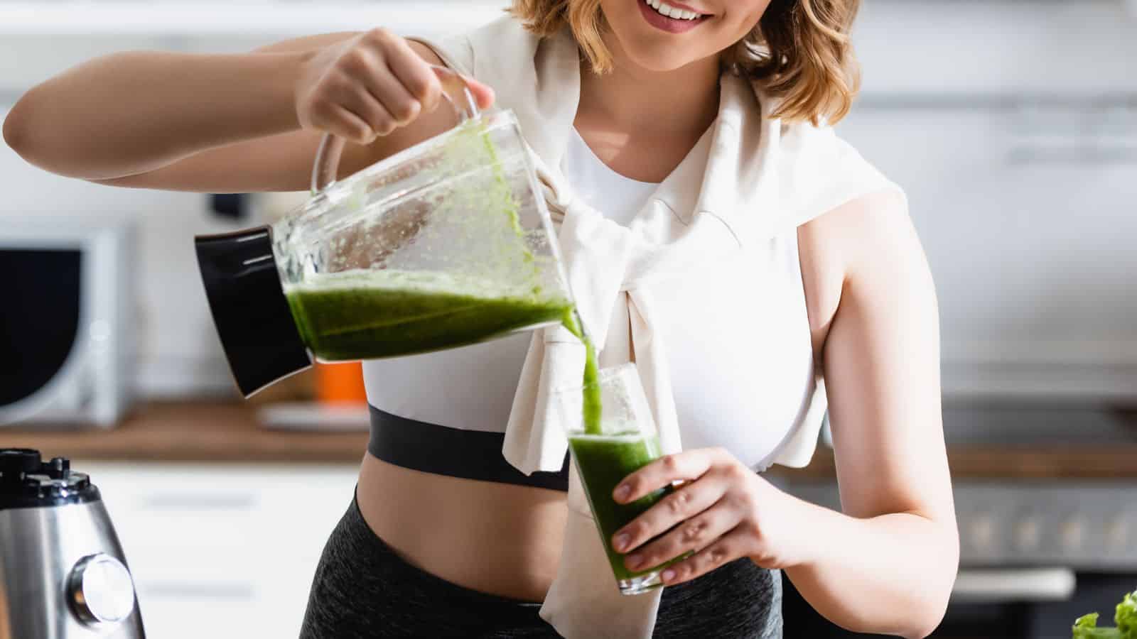 A person in athletic wear is pouring a green smoothie from a blender into a glass. They are standing in a kitchen environment with a smile on their face. A blender base is visible on the counter.