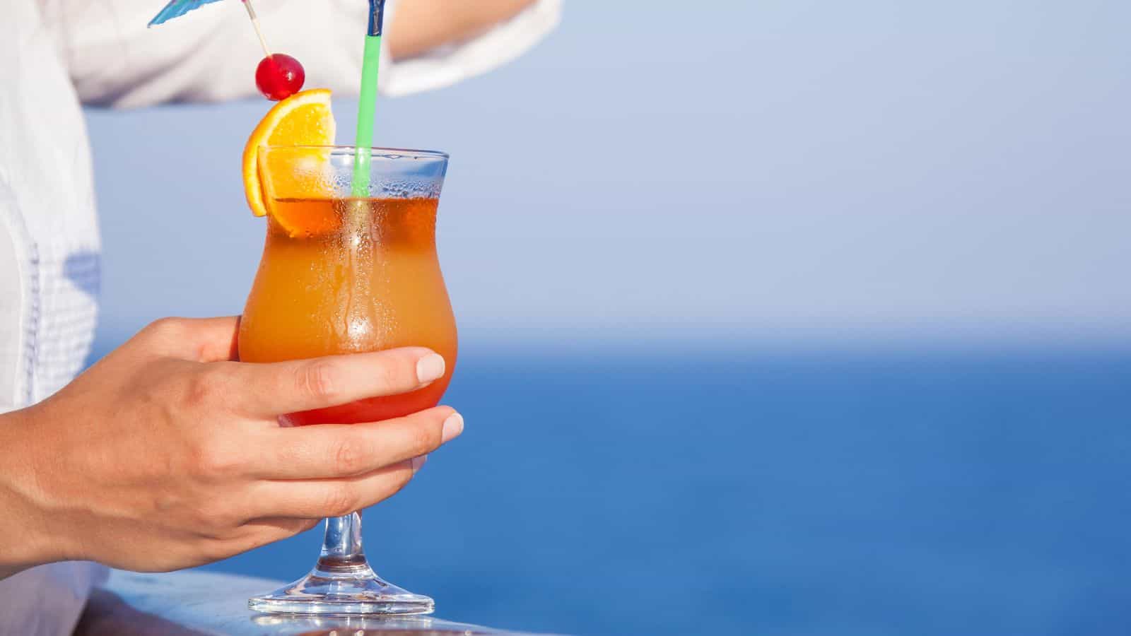 A person holding a tropical cocktail with a garnish of an orange slice and a cherry on a stick. The drink is in a glass with a green straw and blue paper umbrella. The background shows a clear blue sky and sea.