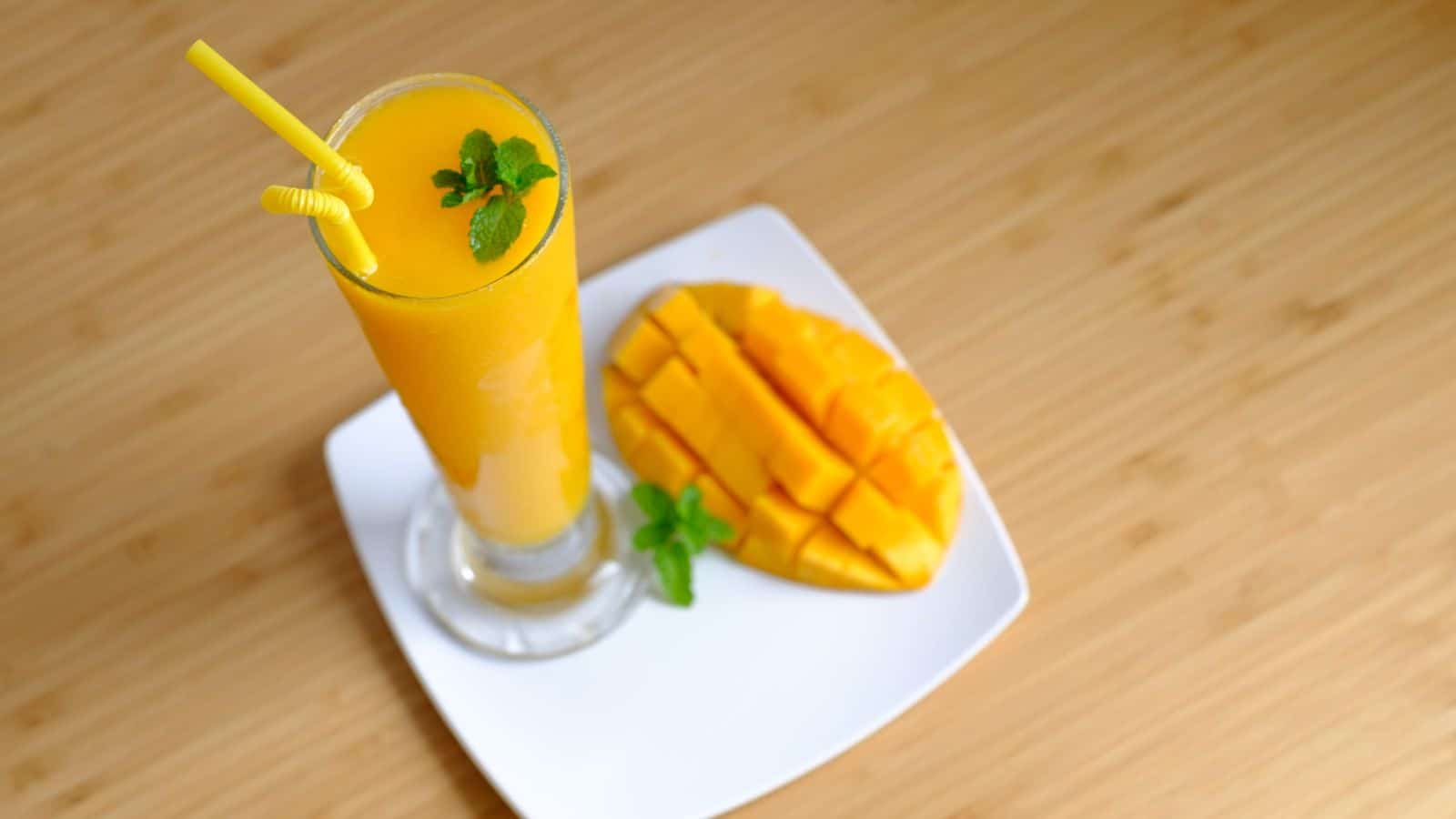 A tall glass of mango juice with a yellow straw is garnished with mint leaves. It is placed on a white square plate with a sliced mango beside it. The background shows a wooden surface.