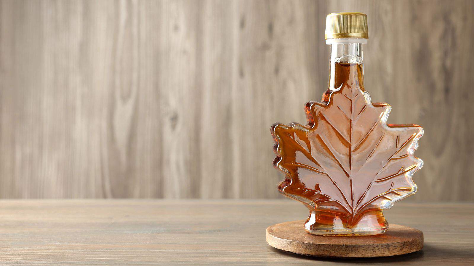 A glass bottle shaped like a maple leaf, filled with amber-colored maple syrup, stands on a round wooden coaster. The background features a textured wood surface.