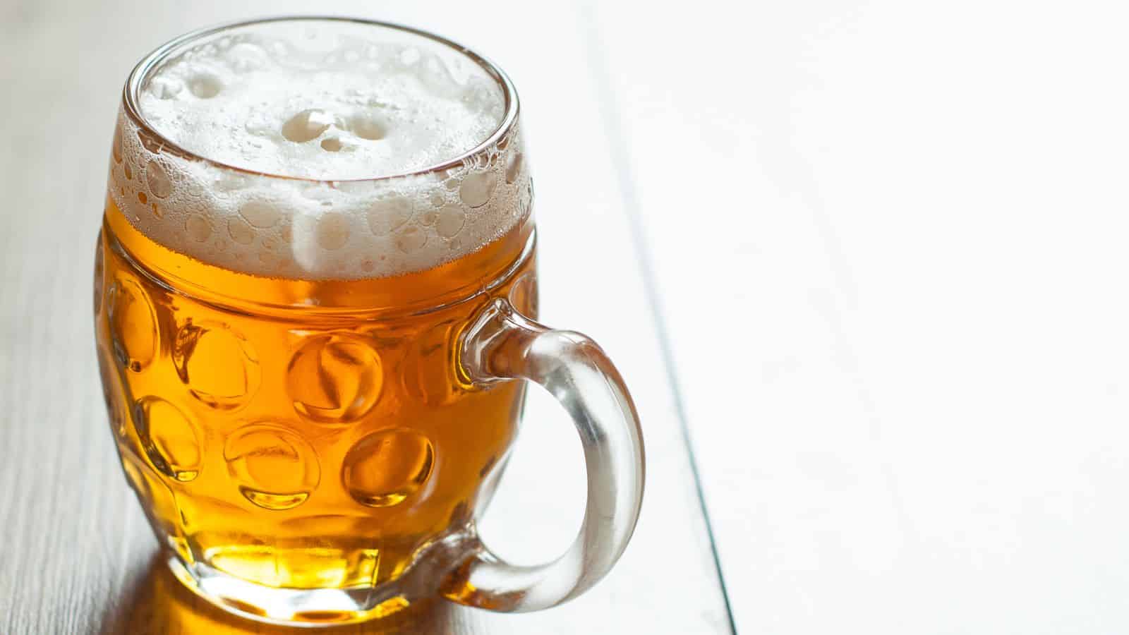A clear glass mug filled with amber-colored beer sits on a wooden surface. The beer has a foamy white head, and the mug features a textured circular pattern with a handle on the right. The background is a pale, wooden texture.
