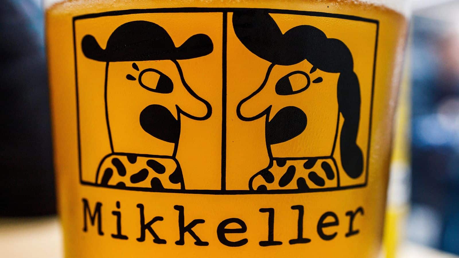 A close-up of a glass filled with beer featuring a Mikkeller logo. The logo shows two stylized faces with cartoonish features, one wearing a hat and the other with long hair, facing each other.