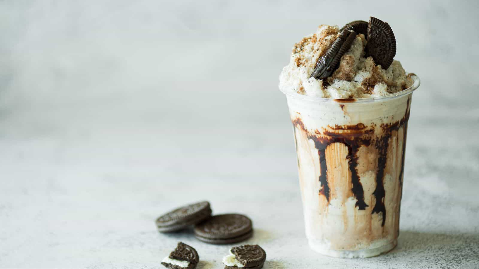 A clear plastic cup filled with cookies and cream milkshake, topped with whipped cream and crushed cookies. Chocolate drizzle is on the inside of the cup. Three Oreo cookies are on the surface nearby. The background is light and blurred.