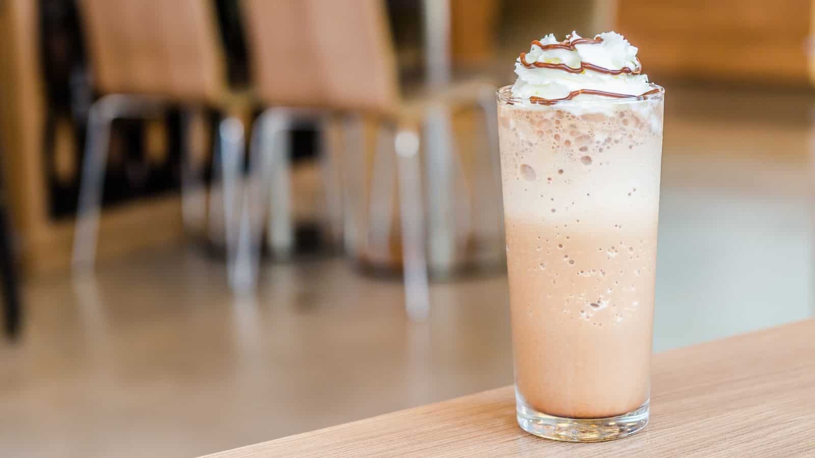 A tall glass of iced coffee topped with whipped cream and drizzled chocolate syrup sits on a wooden table. The background is softly blurred, showing wooden chairs and part of a cafe interior.