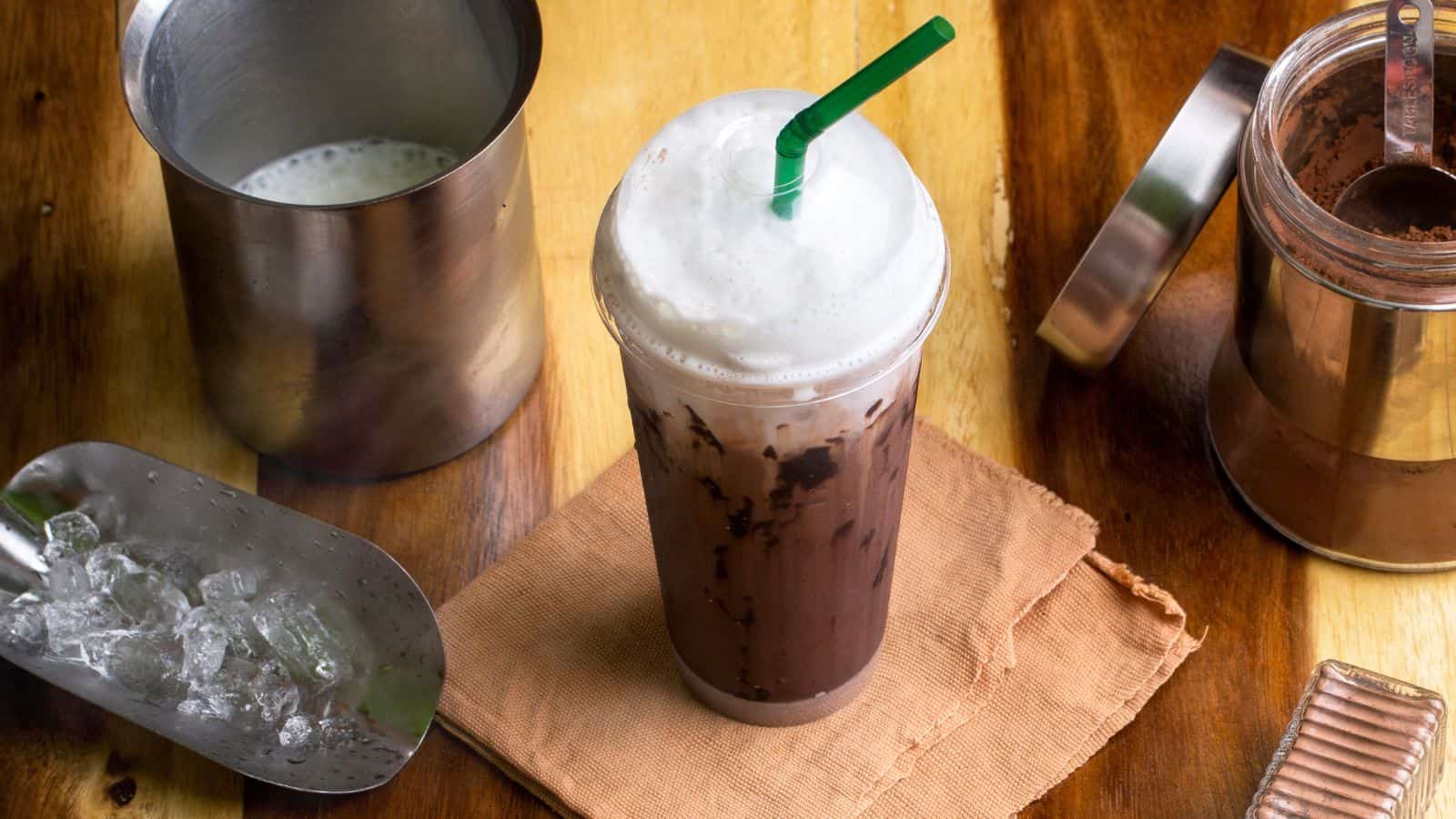 A cold chocolate beverage with whipped cream and a green straw on a napkin, placed on a wooden table. Nearby, there is a metal container with milk, a scoop of ice, and an open jar with cocoa powder.