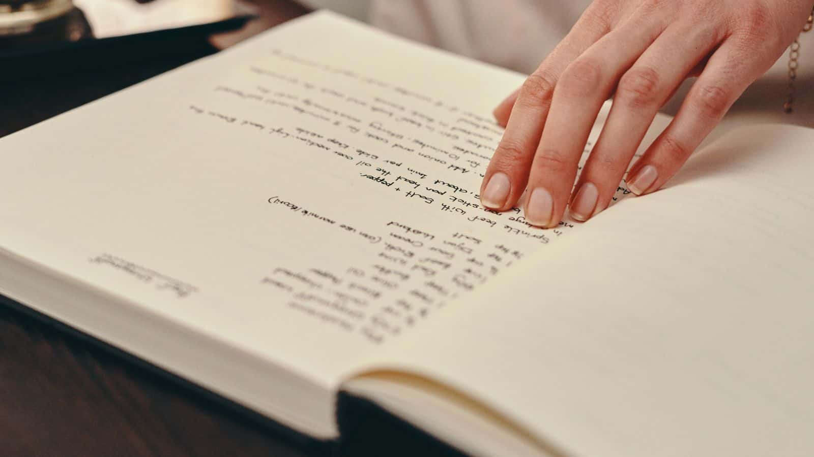 A person's hand gently rests on an open book with handwritten text. The book's pages have neat, evenly spaced writing, and the hand appears to be guiding along the lines. The background is slightly blurred.