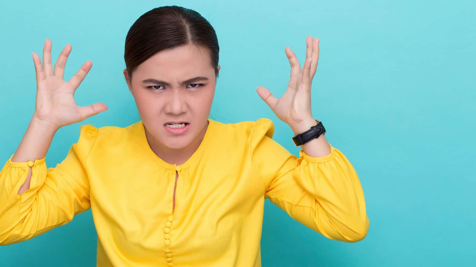 A person with dark hair is wearing a bright yellow shirt and a black watch. They have an angry expression, with their teeth clenched, eyebrows furrowed, and hands raised. The background is a solid light blue.