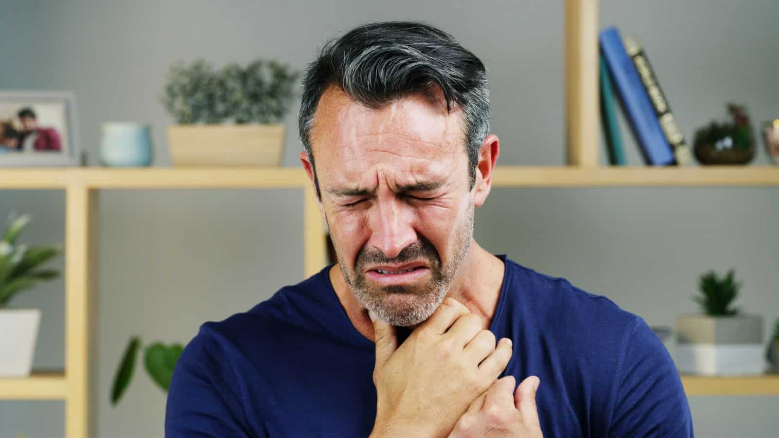 A man with short dark hair and graying temples is in front of wooden shelves. He is wearing a blue shirt and appears to be grimacing, holding his throat with both hands. There are small plants and books on the shelves behind him.