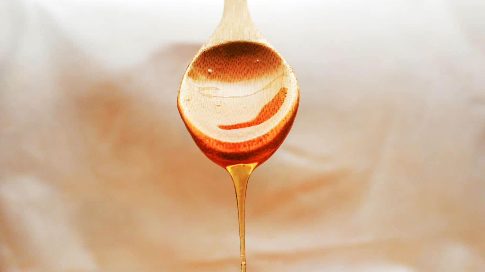 A wooden spoon holds honey, with some dripping off the edge. The background is softly blurred, highlighting the golden color and glossy texture of the honey on the spoon.