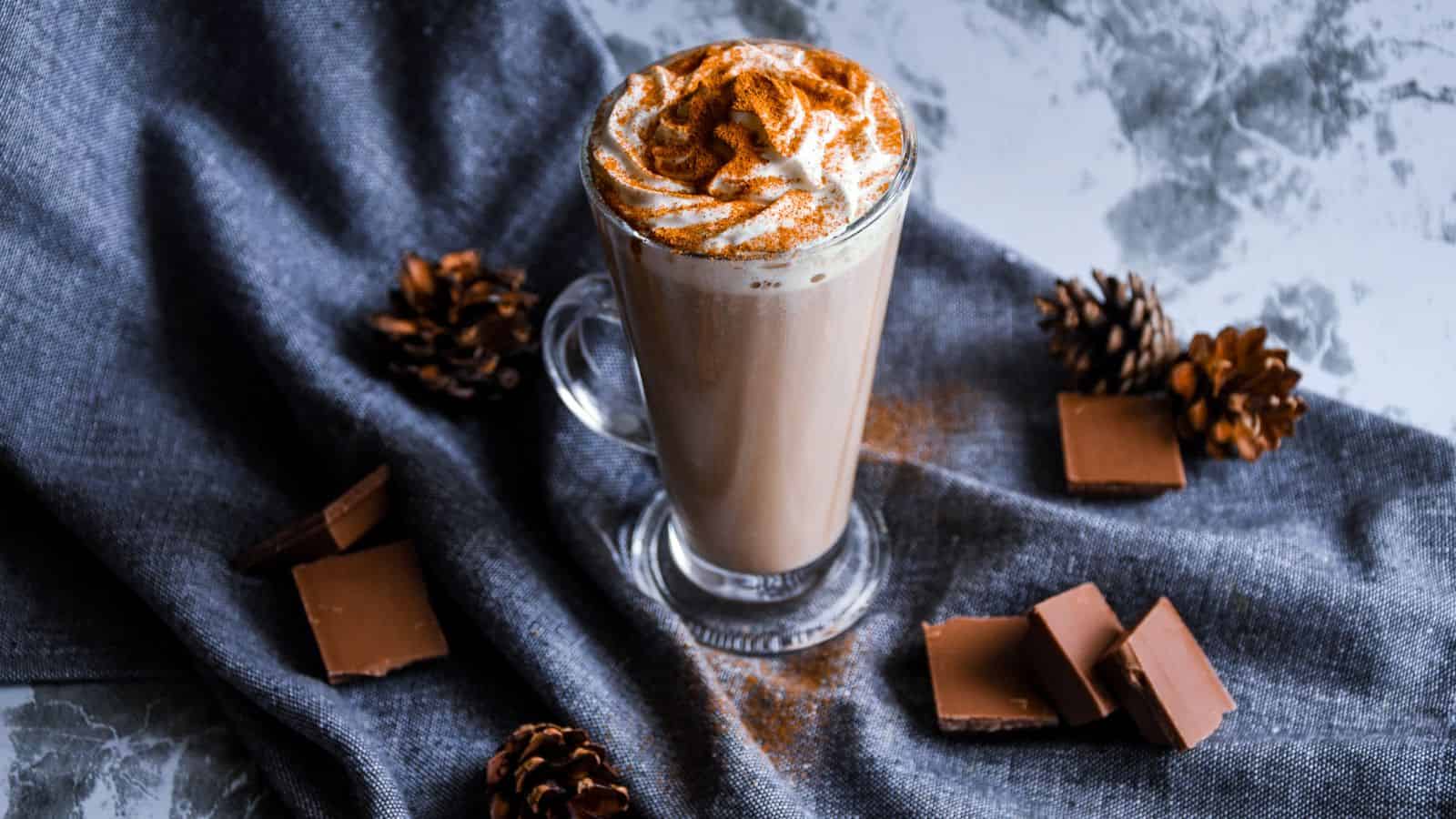 A latte in a tall glass topped with whipped cream and sprinkled with cinnamon sits on a gray fabric. Nearby are pine cones and square pieces of milk chocolate. The setup rests on a textured gray surface.