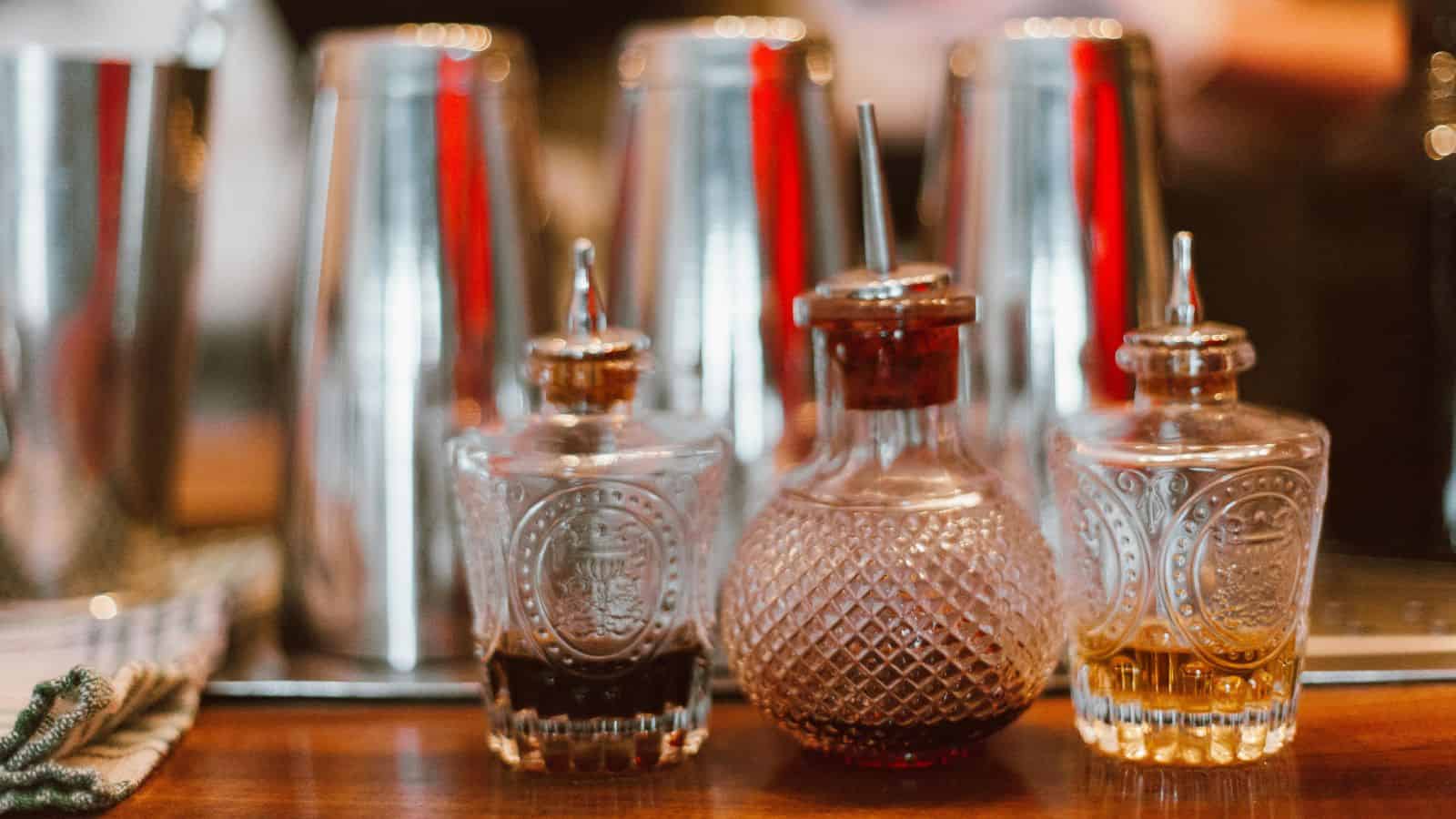 Three glass bottles with metallic pour spouts are on a wooden bar counter. Each bottle contains different colored liquids. In the background, there are several stainless steel shakers lined up, creating a blurred effect.