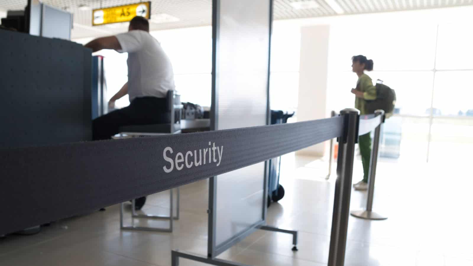 A person sits at a counter in an airport security area, with a black barrier tape labeled "Security" in the foreground. Another individual with a backpack walks in the background. The setting appears to be well-lit with natural light.