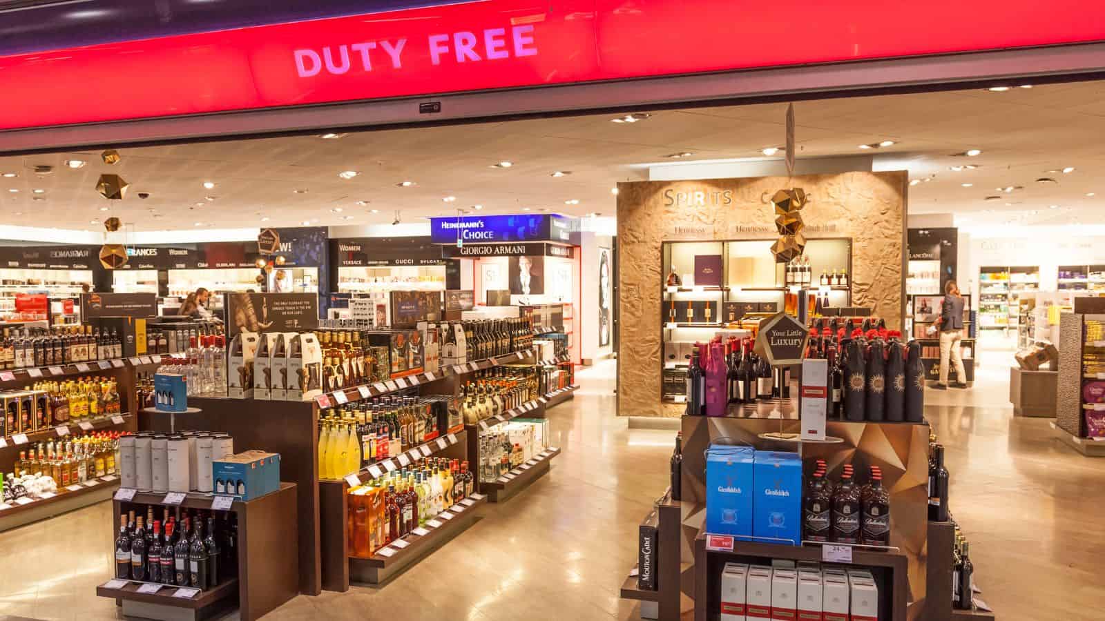 Interior of a duty-free store at an airport. Shelves are stocked with various products including alcohol, chocolates, and perfumes. A red sign above reads "DUTY FREE." The store is brightly lit and organized.