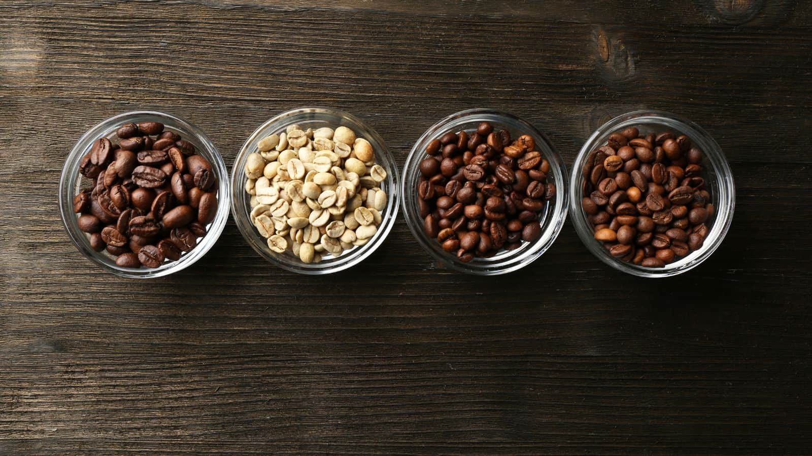 Four glass bowls containing different types of coffee beans are lined up on a wooden surface. From left to right: dark roasted, raw green, medium roasted, and light roasted coffee beans.