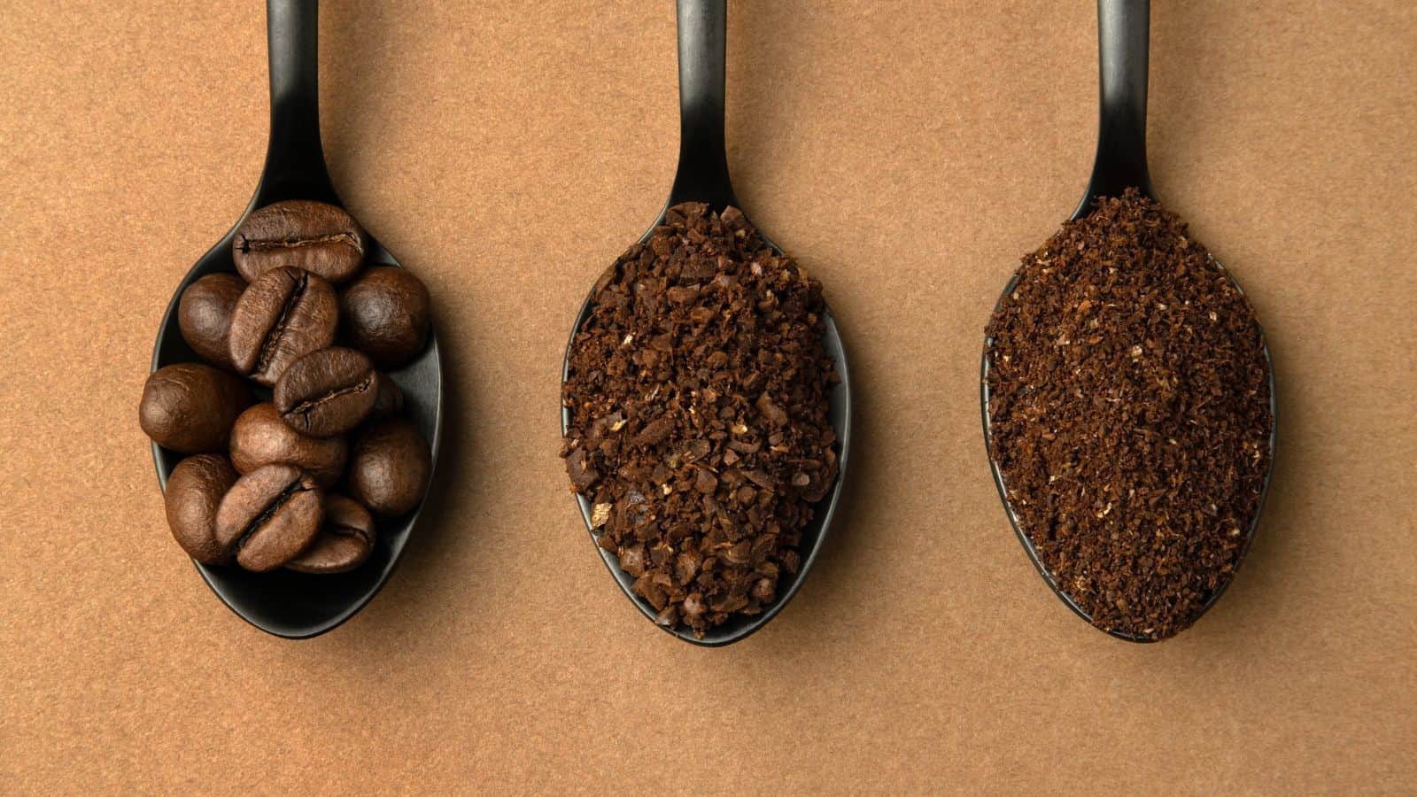 Three black spoons are arranged in a row on a brown surface. The left spoon holds whole coffee beans, the middle spoon has coarsely ground coffee, and the right spoon contains finely ground coffee.