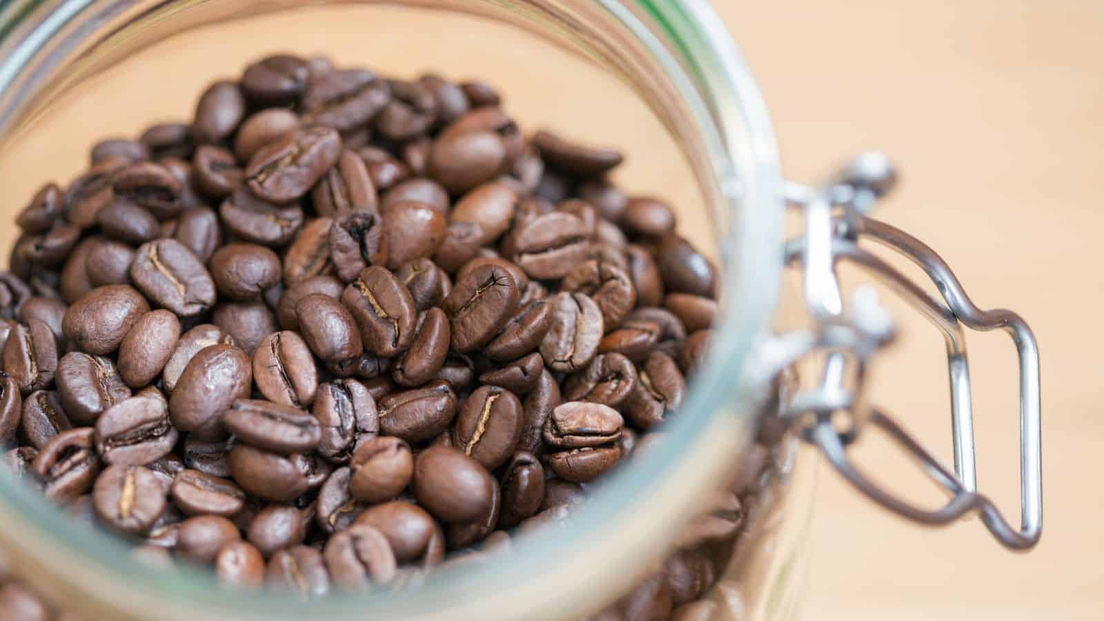 A glass jar filled with whole coffee beans. The jar has a metal clasp and is open, revealing a close-up view of the dark brown beans. The background is a soft beige color, slightly blurred.