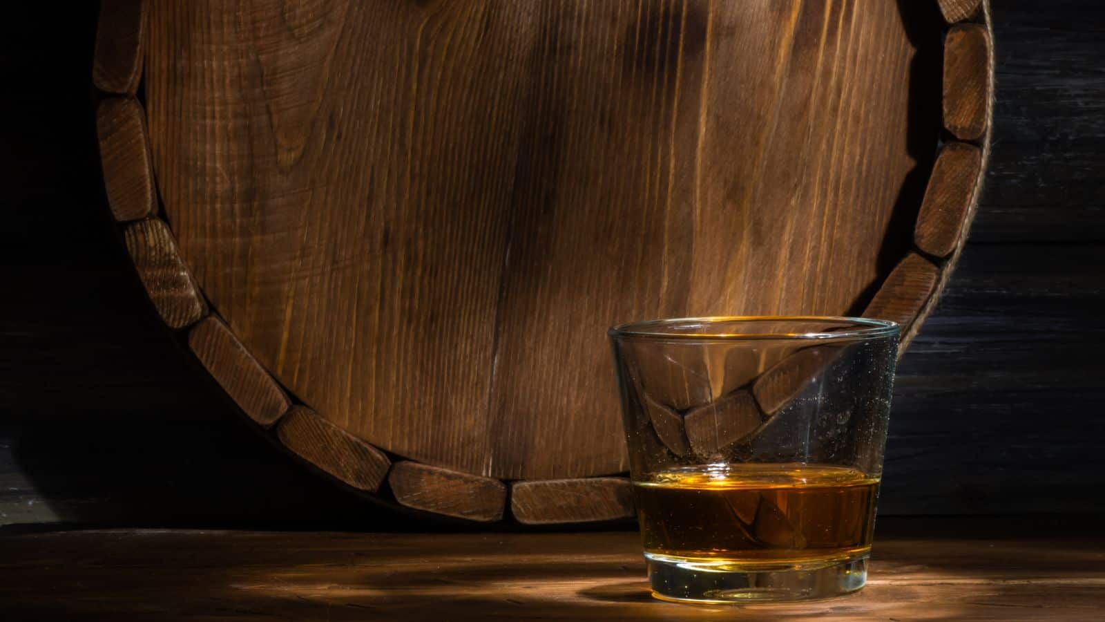 A glass filled with amber liquid sits on a wooden surface, illuminated by light. Behind the glass, there is a large, round wooden barrel with visible grain and texture. The scene conveys a rustic and warm atmosphere.
