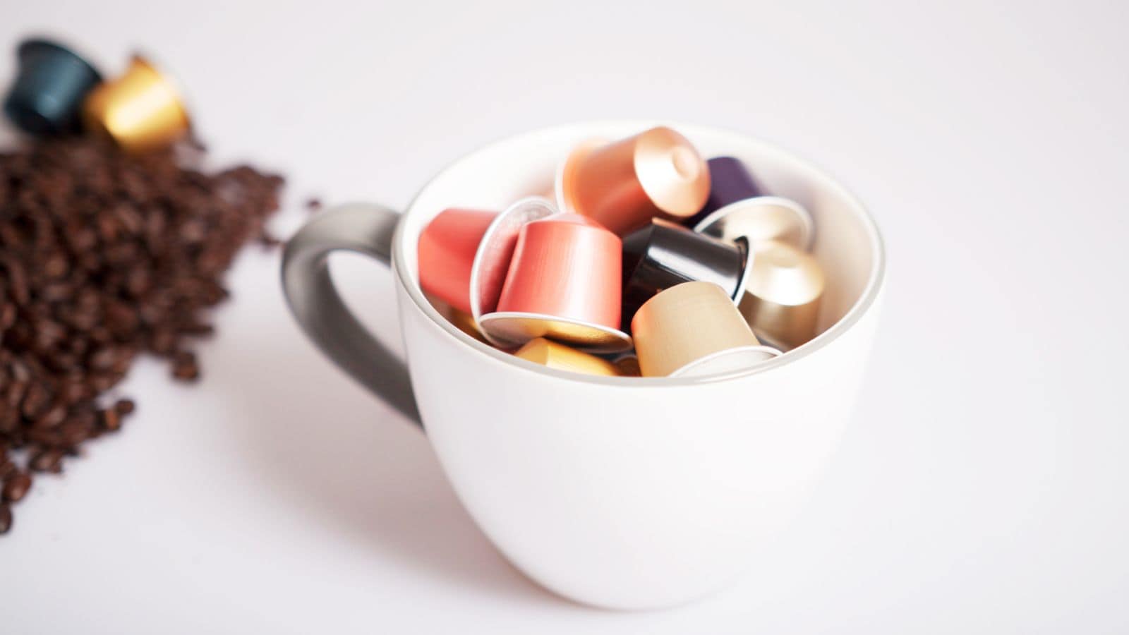 A white cup filled with various colored coffee pods sits on a white surface. To the left of the cup, there is a small pile of coffee beans. The pods are metallic and cylindrical, each a different color.